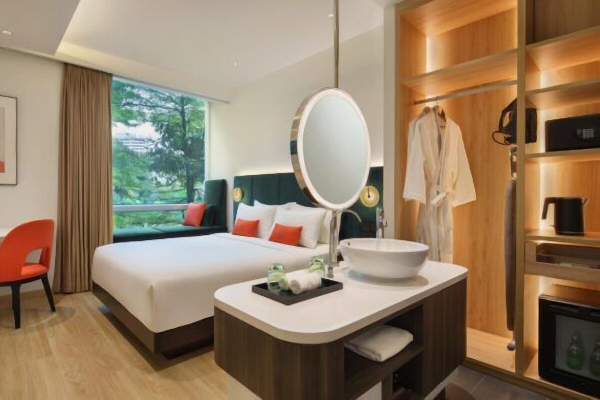 A hotel room with a bed, a desk, a chair, a bathroom with a sink and mirror, and a view of greenery.
