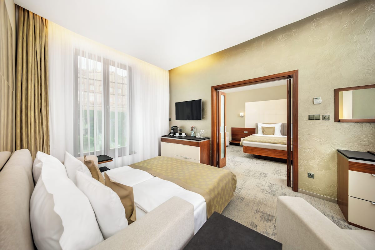 A hotel room with a bed, bedside table, and a desk with a chair. There is a window with curtains, a flat-screen TV mounted on the wall, and a mirror above the desk.