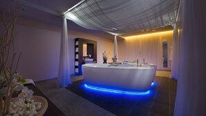 A modern spa room with a large bathtub, blue ambient lighting, and a serene decor including white flowers and a wooden bench. - Wellness Hotel Diamant (Hluboka nad Vltavou)