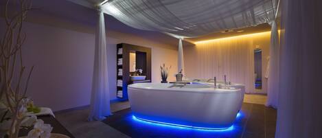 Un spa moderno con una bañera grande, iluminación ambiental azul y una decoración serena que incluye flores blancas y un banco de madera.