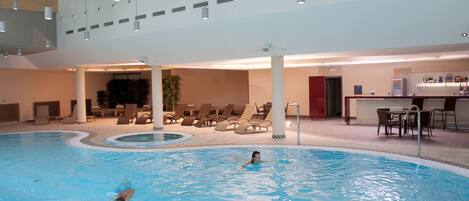 Una piscina interior con una persona nadando, sillas de descanso y una zona de bar.