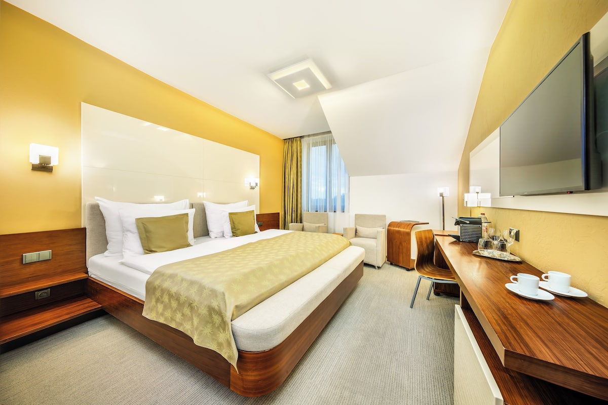 A hotel room with a large bed, a wooden desk, a flat-screen TV, and a view of the city through the window.