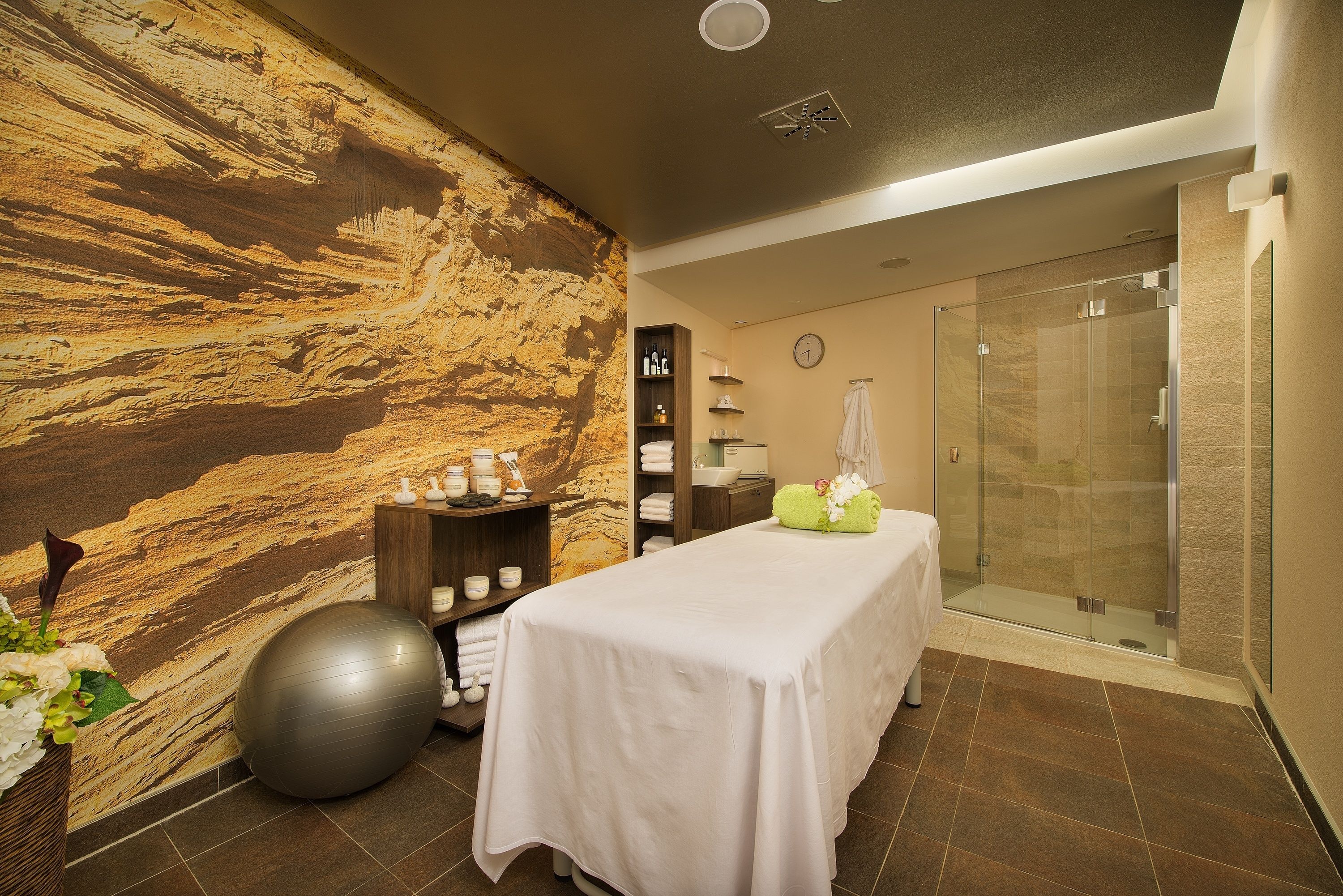 Un cuarto de spa con una camilla de masaje, una pelota grande de ejercicios, una pared con textura, una cabina de ducha de vidrio y un estante con artículos de tocador.