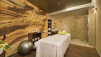 Un cuarto de spa con una camilla de masaje, una pelota grande de ejercicios, una pared con textura, una cabina de ducha de vidrio y un estante con artículos de tocador.