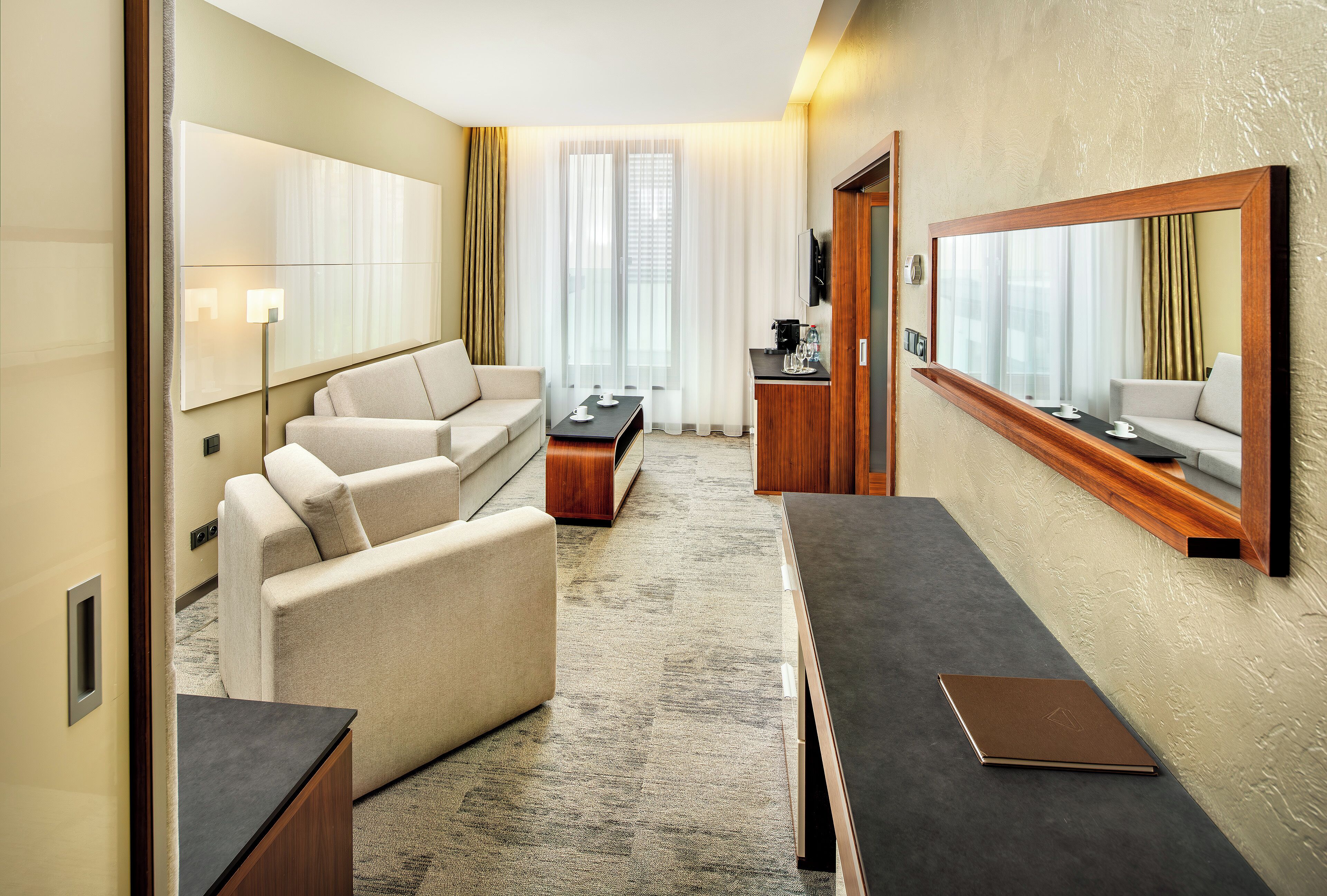 A modern hotel room with a sofa, a coffee table, a desk, and a large mirror.
