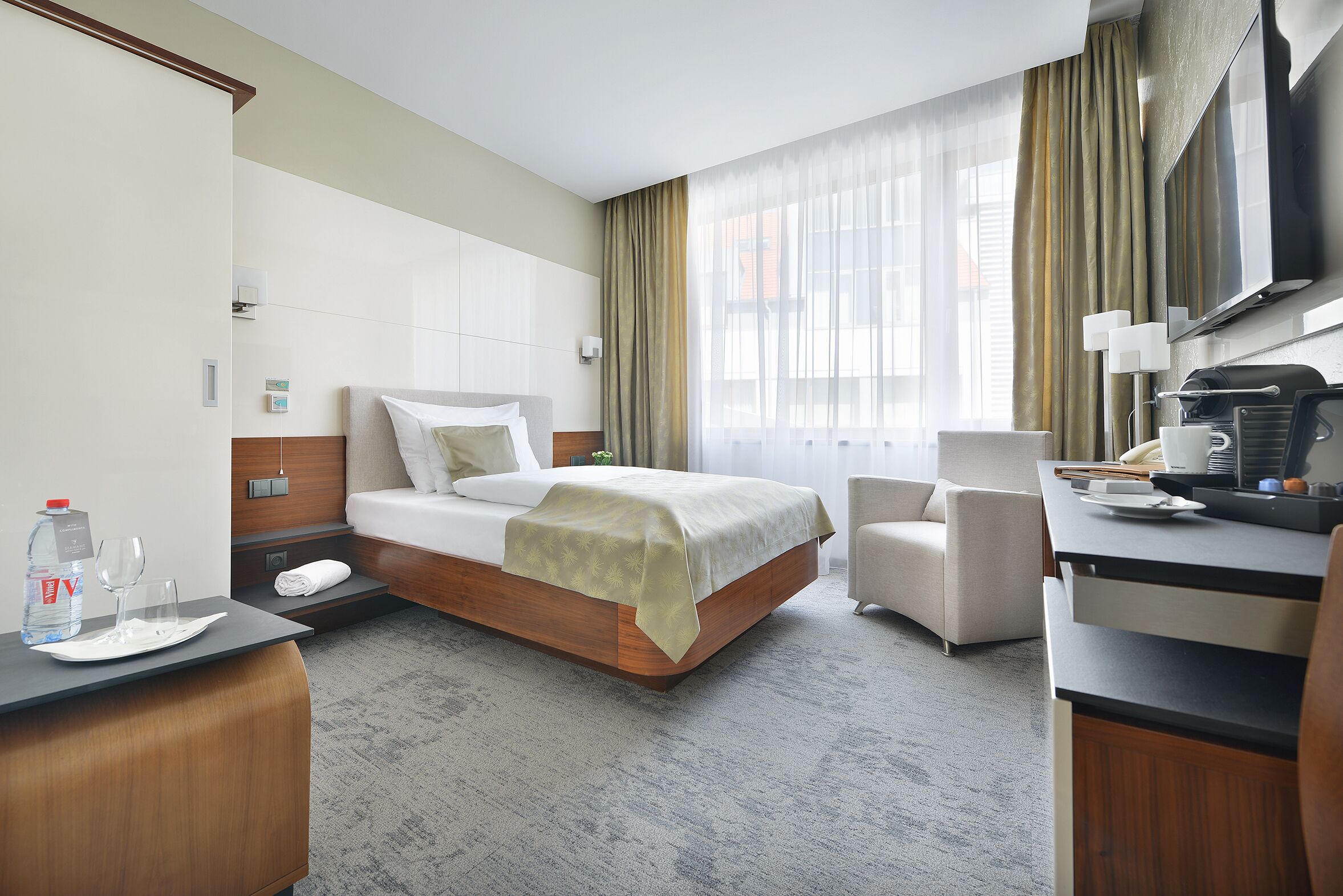 A modern hotel room with a bed, bedside table, chair, desk, and a large window with curtains.