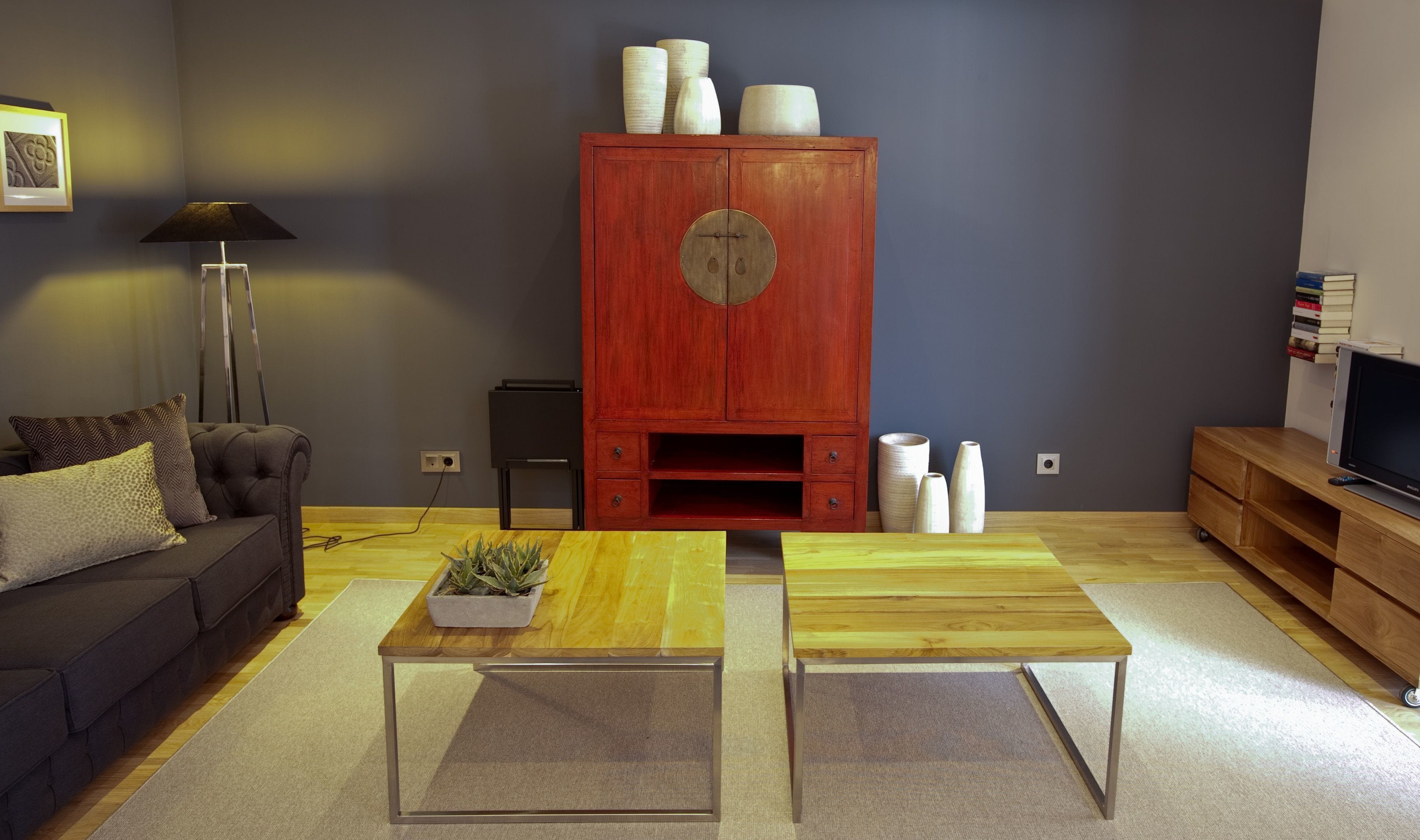 Una sala de estar con un sofá oscuro, dos mesas de centro de madera, un mueble rojo con objetos decorativos y un televisor sobre un soporte de madera.