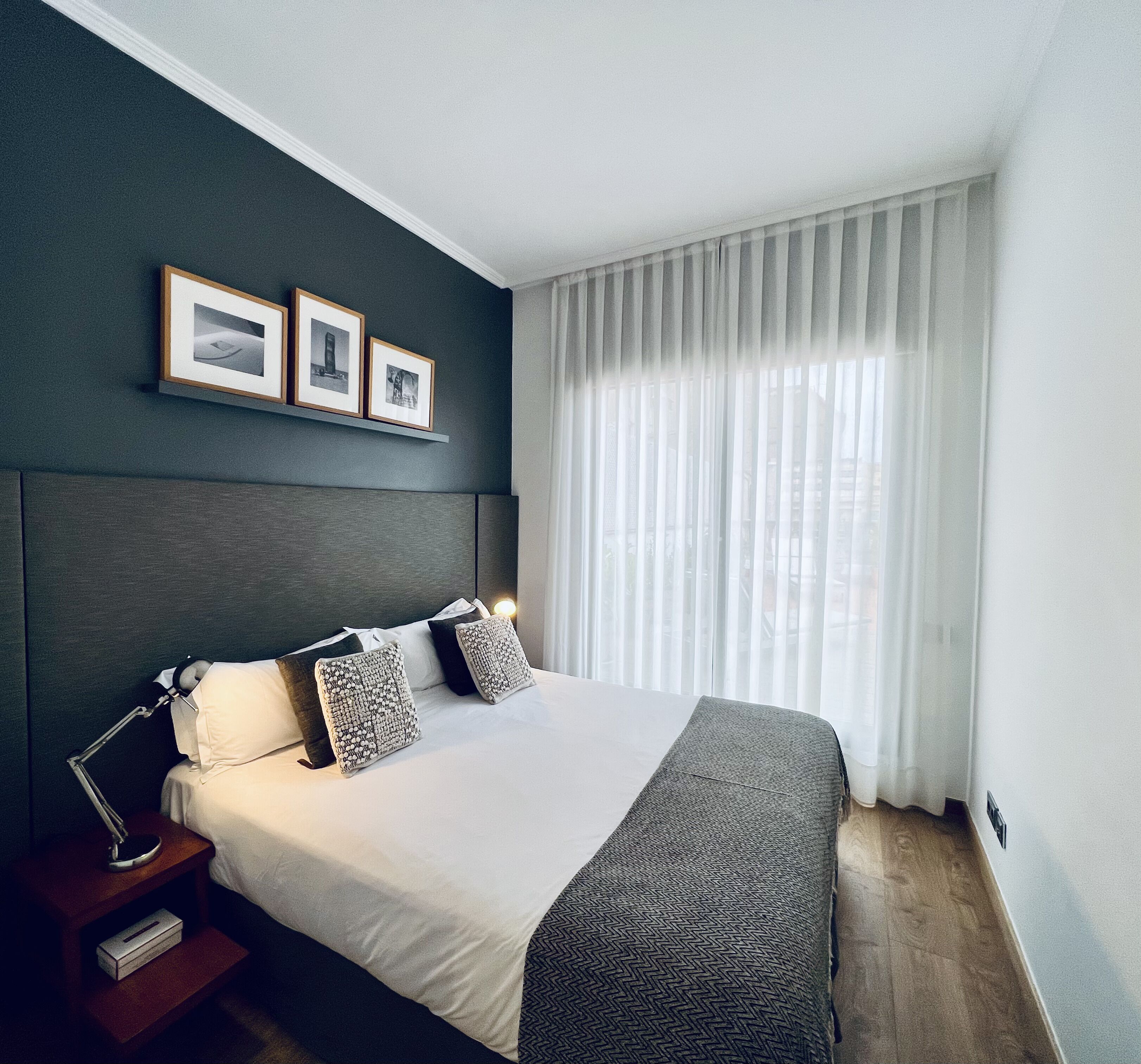 A modern hotel room with a large bed, a bedside table, a lamp, and a window with sheer curtains.
