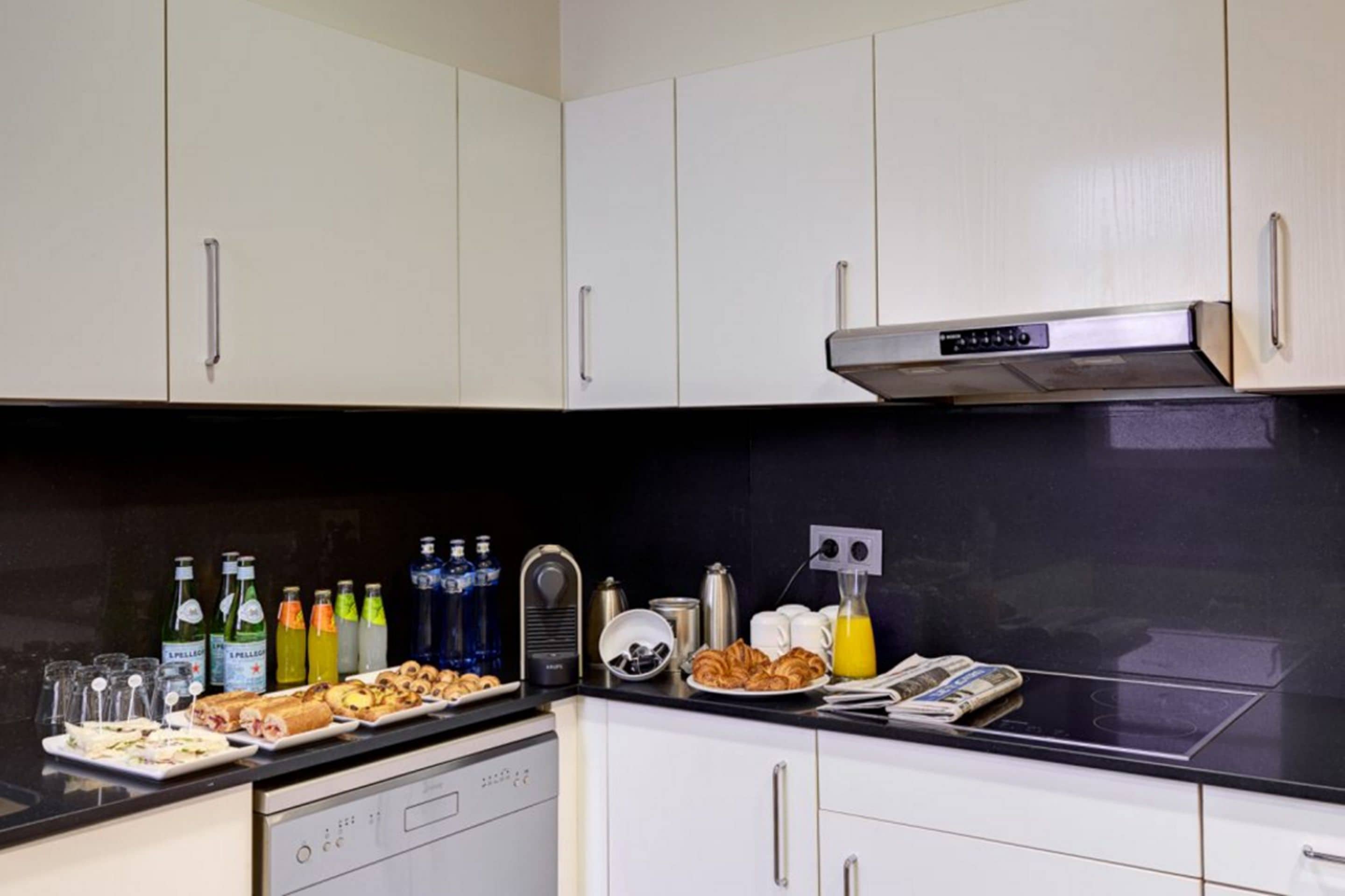 Una cocina con armarios blancos, encimera negra y diversos productos para el desayuno, como cruasanes, jugo y café.