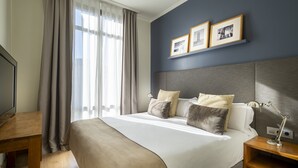 Una cama bien tendida con cabecera, una mesita de noche con lámpara y un televisor a la izquierda.
