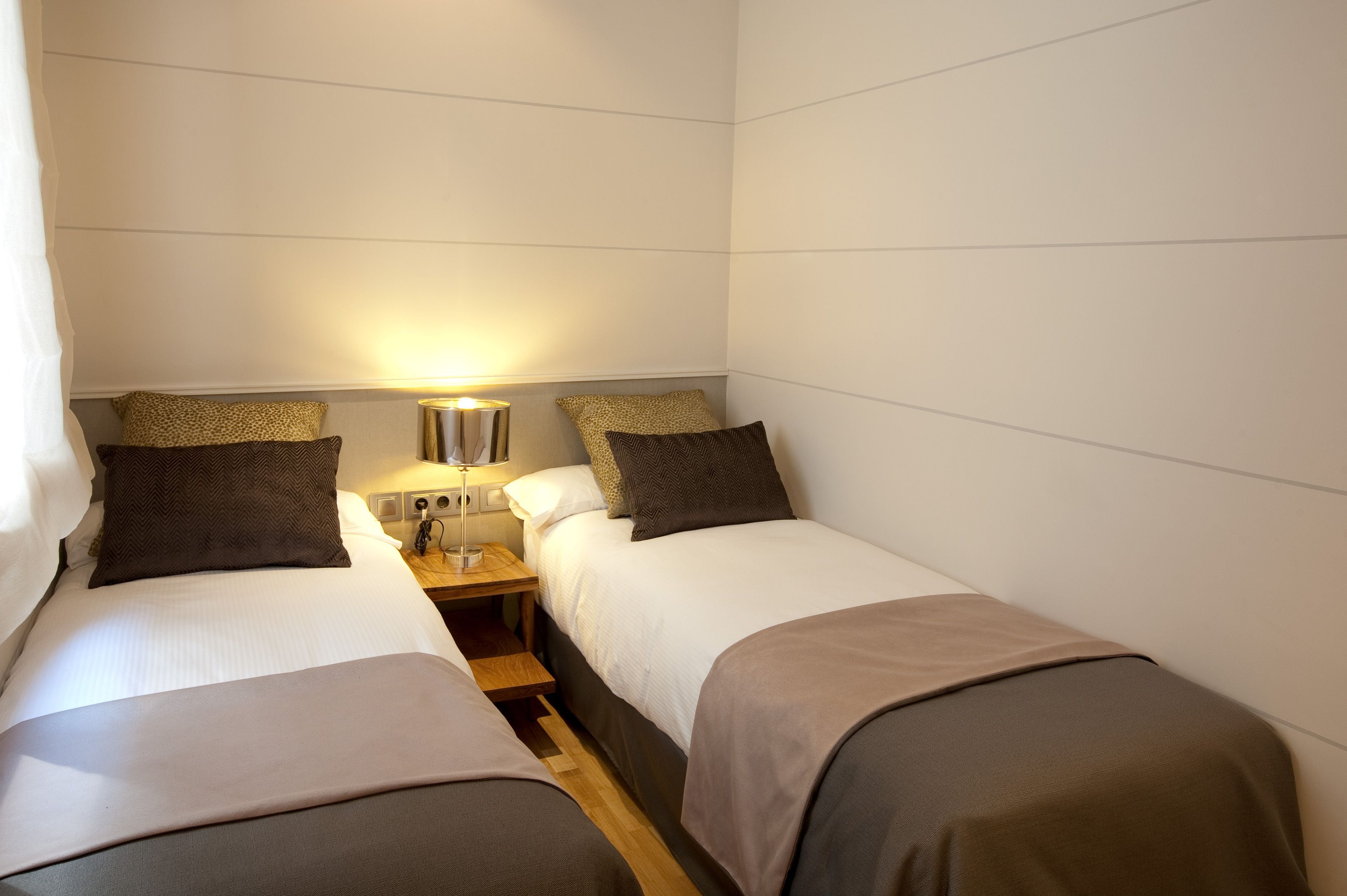 A small, compact hotel room with two single beds, a bedside table with a lamp, and a power outlet.