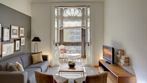Una sala moderna con sofá, mesa de centro y televisor. Hay cuadros en la pared y un ventanal con vistas a edificios.