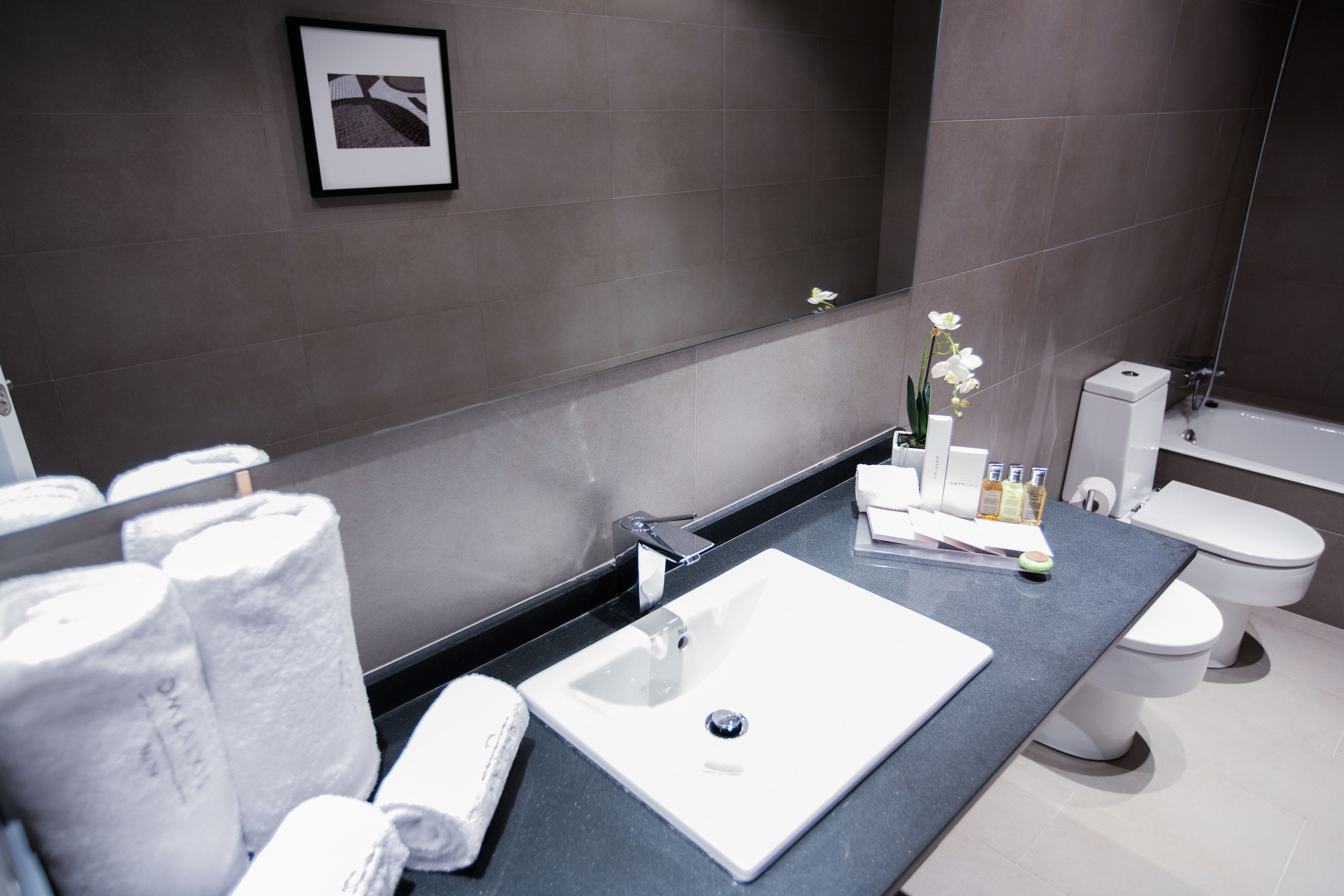 Ein modernes Badezimmer mit einem weißen Waschbecken, einem großen Spiegel und einer Toilette. Auf der Ablage liegen Handtücher und Pflegeartikel.