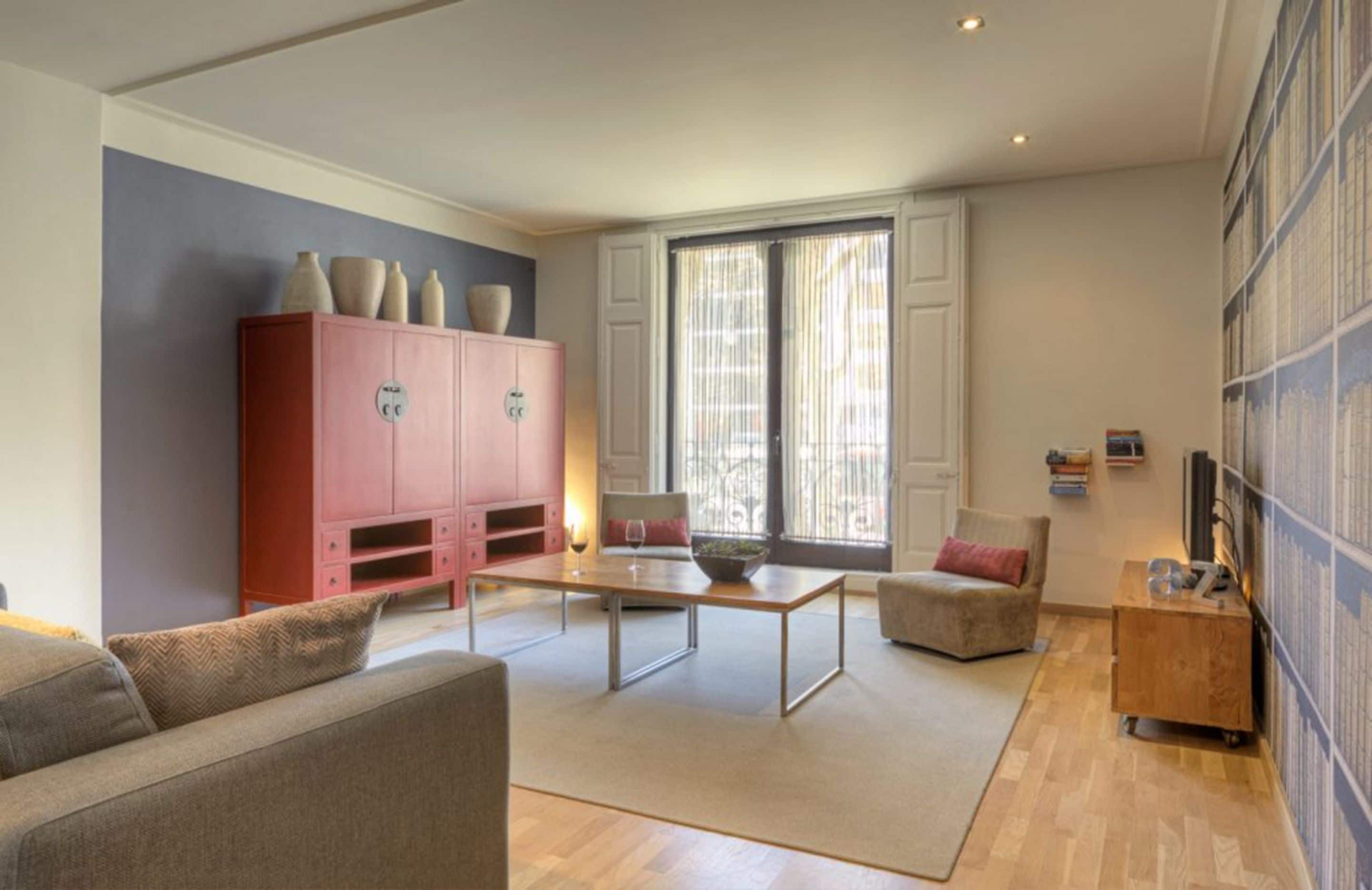 A modern living room with a sofa, a coffee table, a pink cabinet with decorative items, and a large window with sheer curtains.
