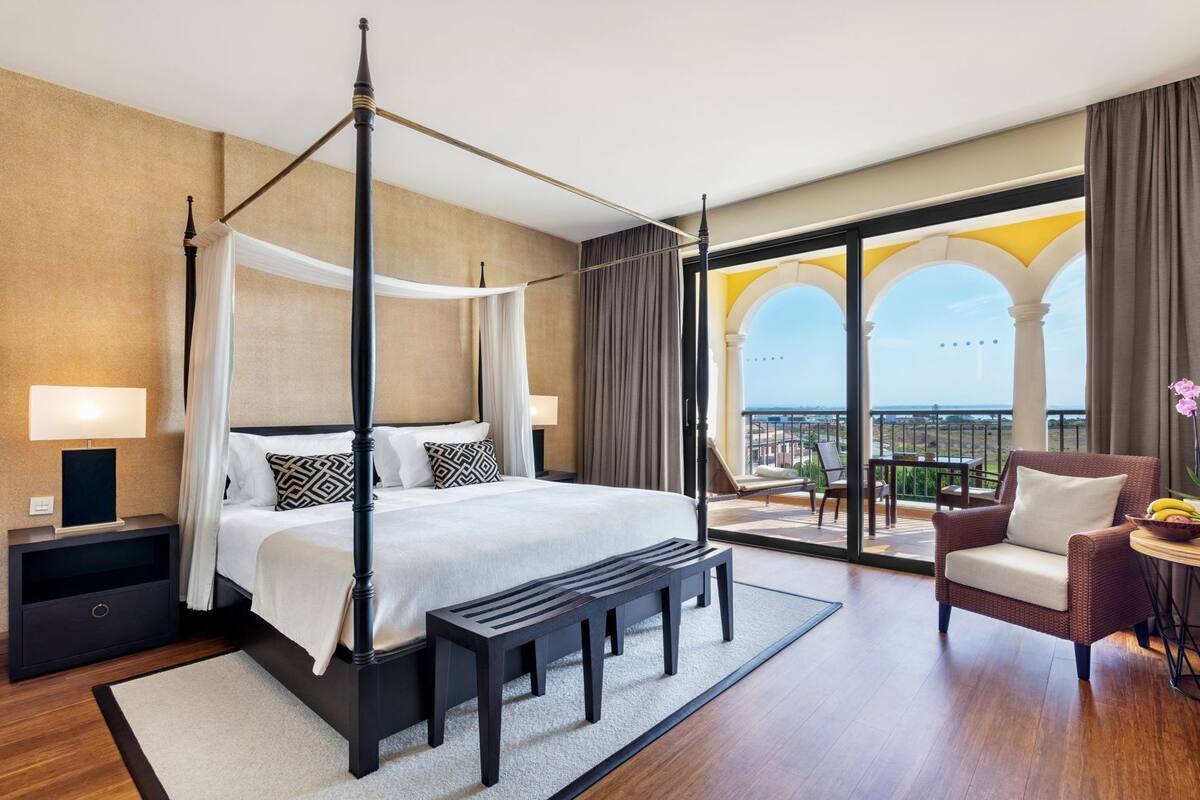 Una cama con dosel, una mesita de noche con lámpara, un banco, una silla y vistas al balcón con mobiliario exterior.