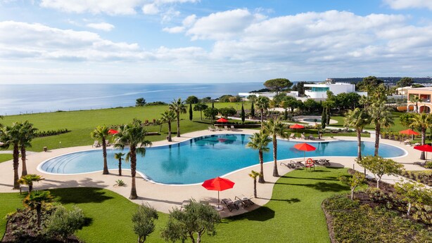 A large swimming pool with multiple red umbrellas, surrounded by palm trees and a view of the sea. - Cascade Wellness Resort Lagos Algarve by Domes (Lagos)