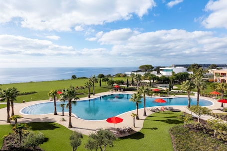 A large swimming pool with multiple red umbrellas, surrounded by palm trees and a view of the sea.