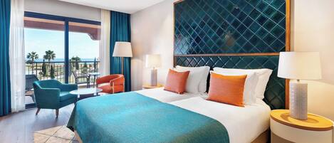 A hotel room with a large bed, two bedside tables, a chair, a balcony with a view of palm trees and the sea.