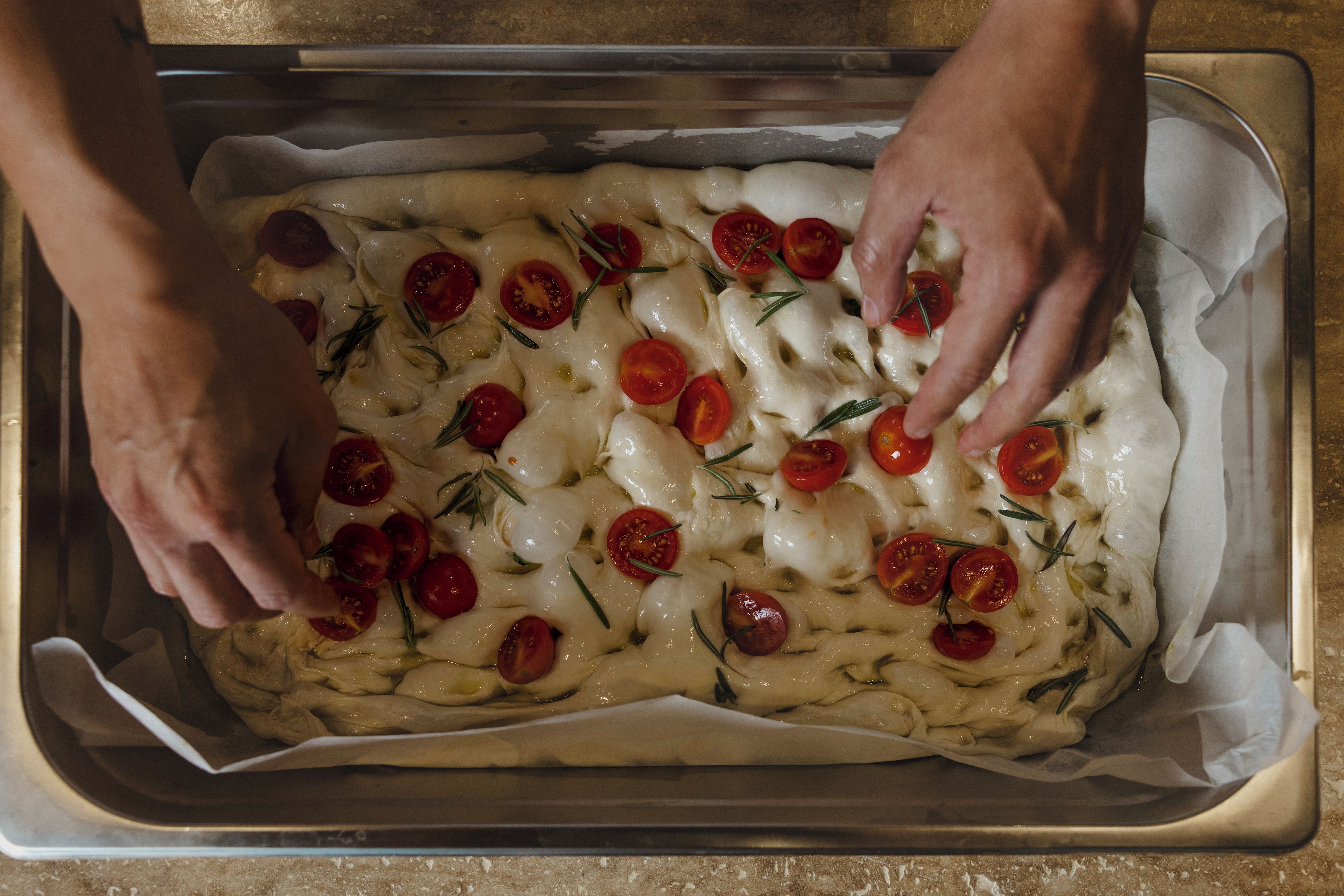 Eine Person legt Kirschtomaten und Rosmarin auf ein Backblech.