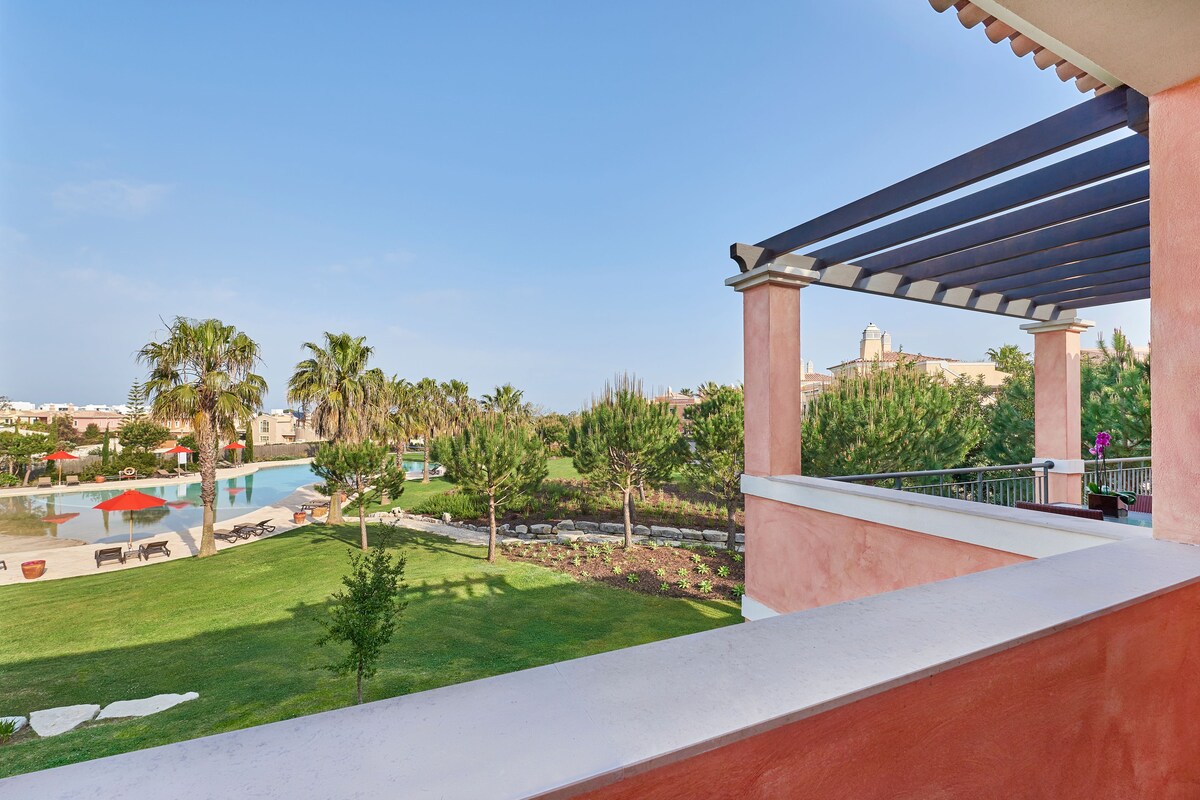 A balcony with a view of a swimming pool, palm trees, and a distant building.