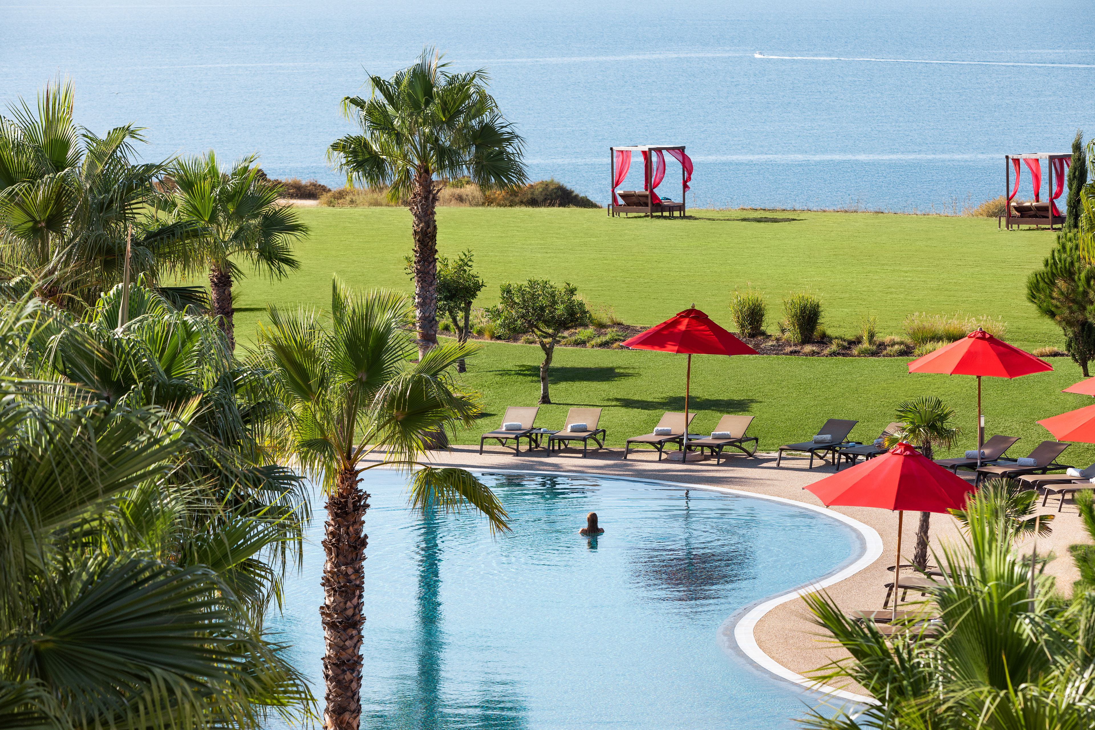 Una piscina con una persona nadando, rodeada de palmeras y sombrillas rojas, con vista al mar y un área verde con sillones de descanso.