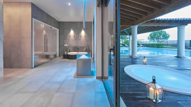 A modern spa area with a glass-enclosed sauna, a seating area with a massage table, and a view of a pool and outdoor space.