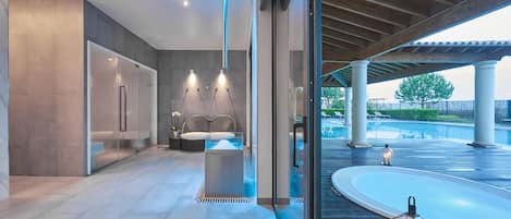 Un moderno espacio de spa con sauna de cristal, una zona de estar con camilla de masaje y vistas a una piscina y un espacio exterior.