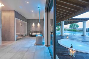 A modern spa area with a glass-enclosed sauna, a seating area with a massage table, and a view of a pool and outdoor space.
