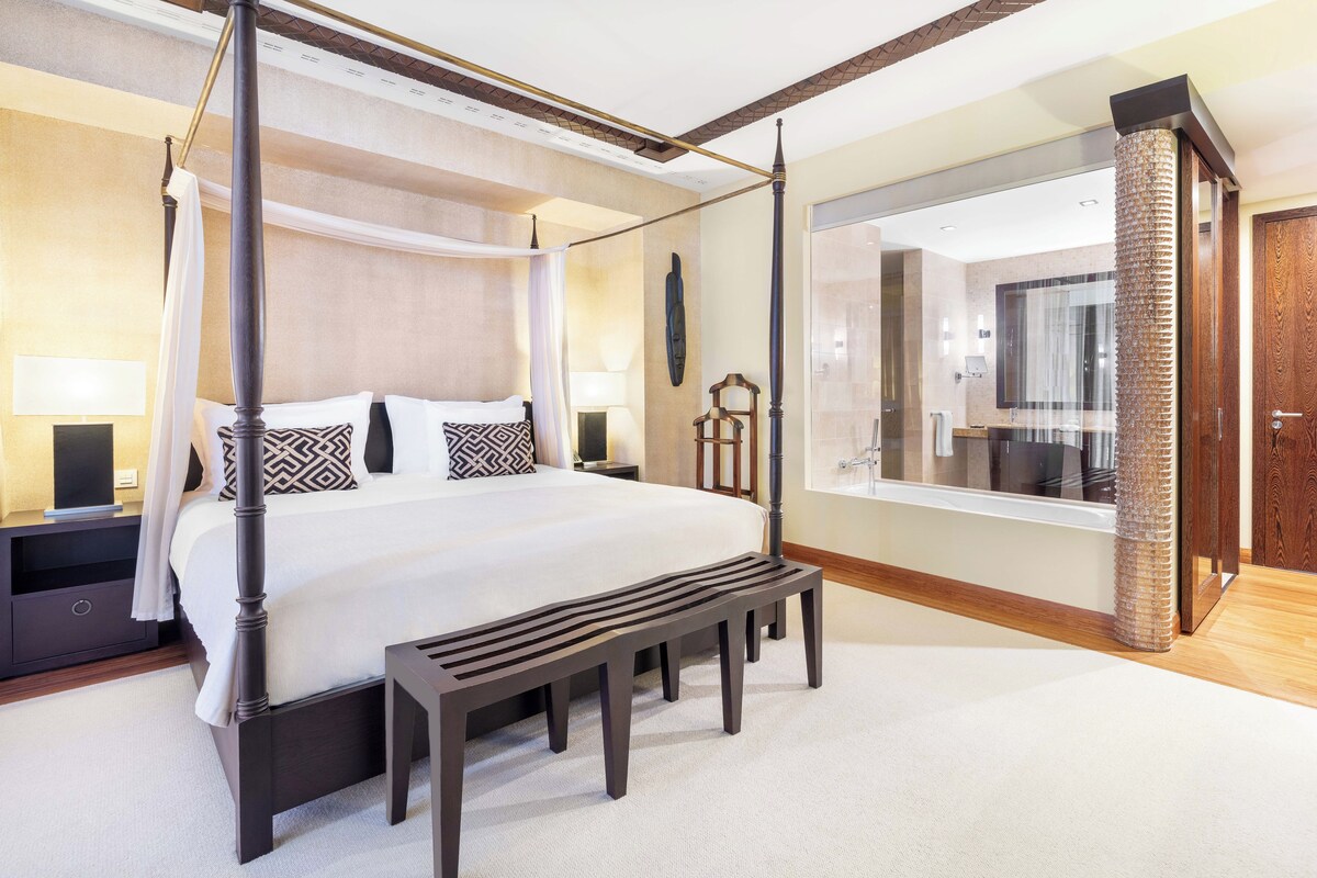 A four-poster bed with a bench in front, a nightstand with a lamp, and a large mirror reflecting the bathroom.