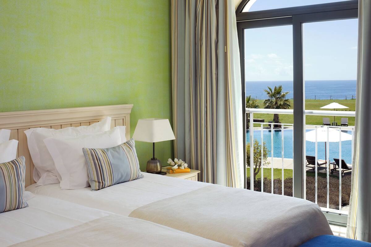 Habitación de hotel con una cama, una mesita de noche, una lámpara y vistas a una piscina y al océano.