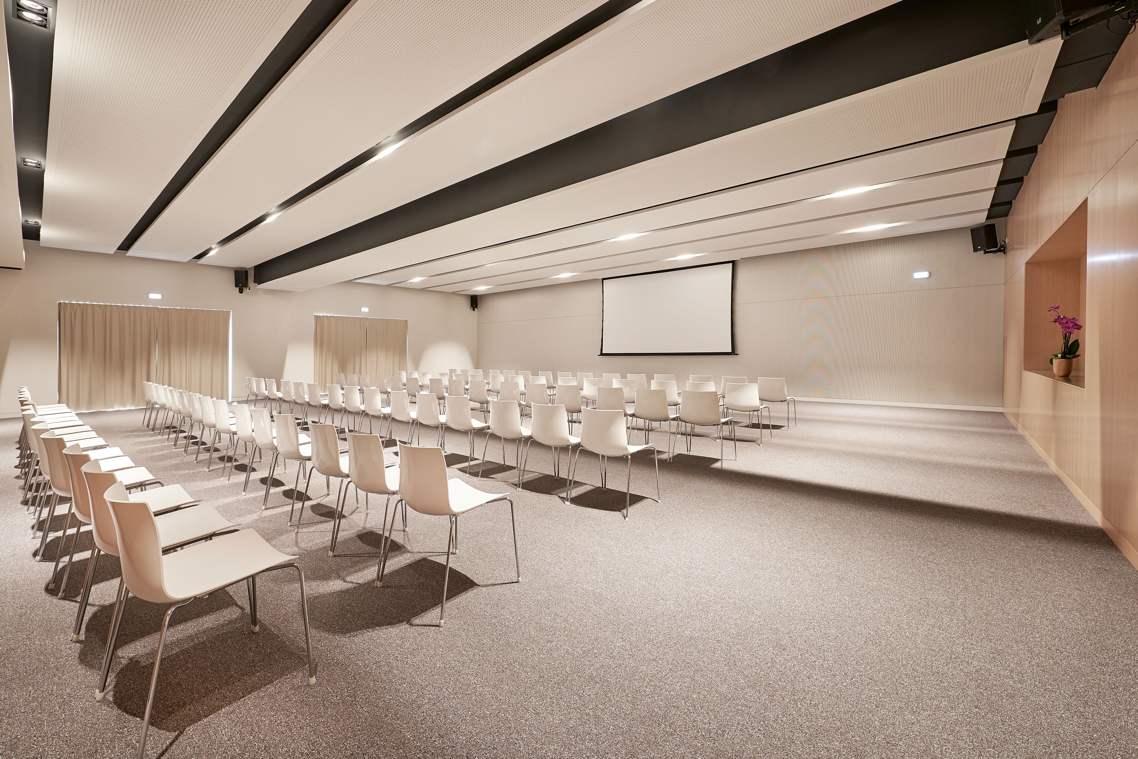 Une salle de conférence avec des rangées de chaises, un écran de projection et un sol recouvert de moquette.