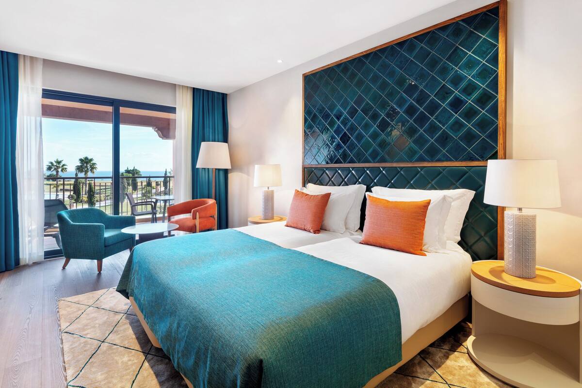 Habitación de hotel con una cama grande, dos mesitas de noche, una silla y un balcón con vistas a palmeras y al mar.
