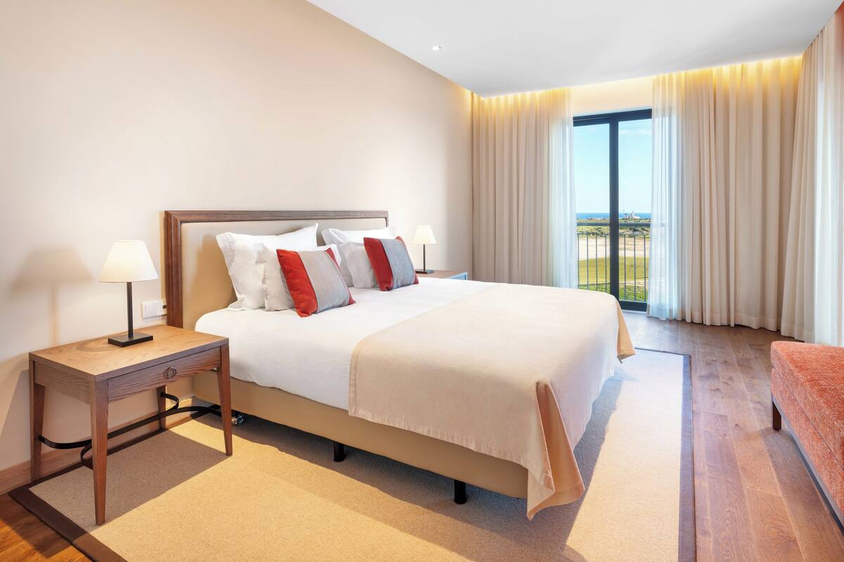 Habitación de hotel con una cama grande, una mesita de noche, una lámpara y vistas al exterior a través de un ventanal amplio.