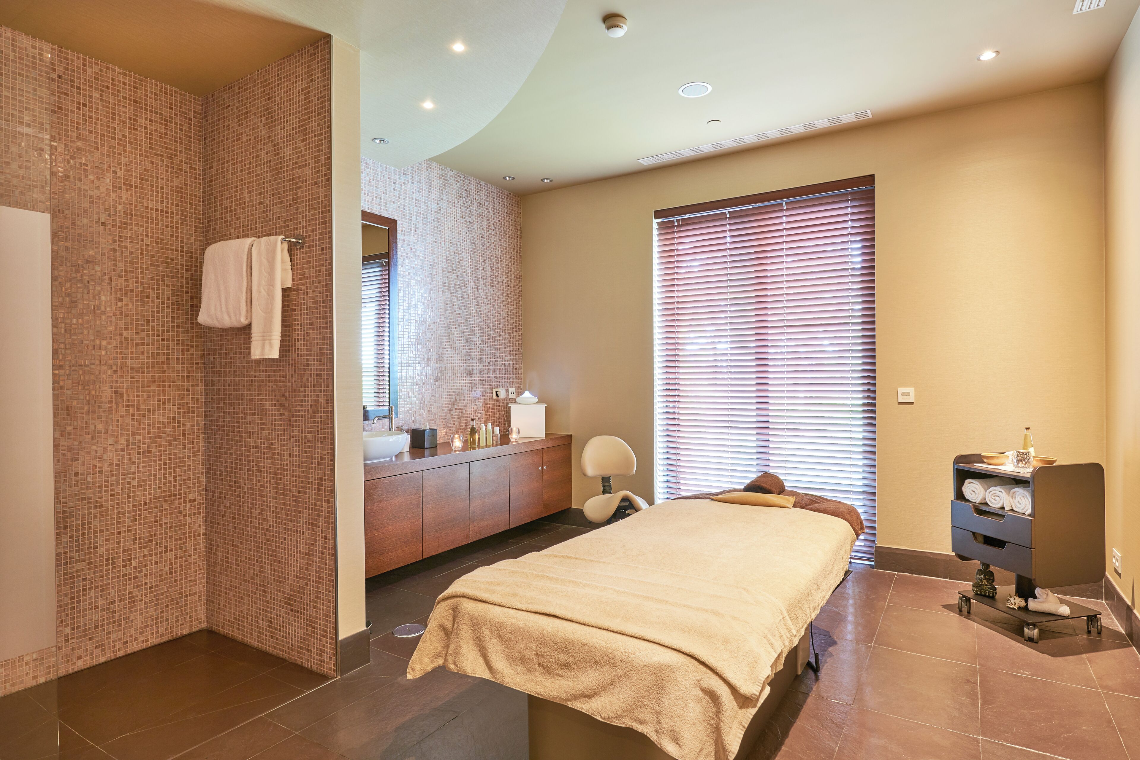 A modern hotel room with a large bed, a bathroom with a tiled shower wall, a vanity with toiletries, and a window with blinds.