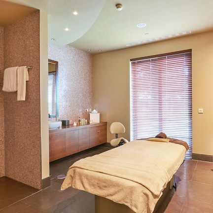 A modern hotel room with a large bed, a bathroom with a tiled shower wall, a vanity with toiletries, and a window with blinds.