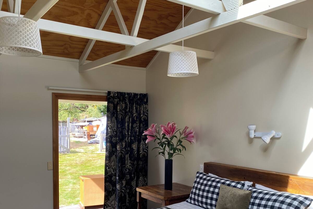 A bedroom with a checkered bedspread, a wooden headboard, a nightstand with a vase of flowers, and a view of the outdoors through a glass door.