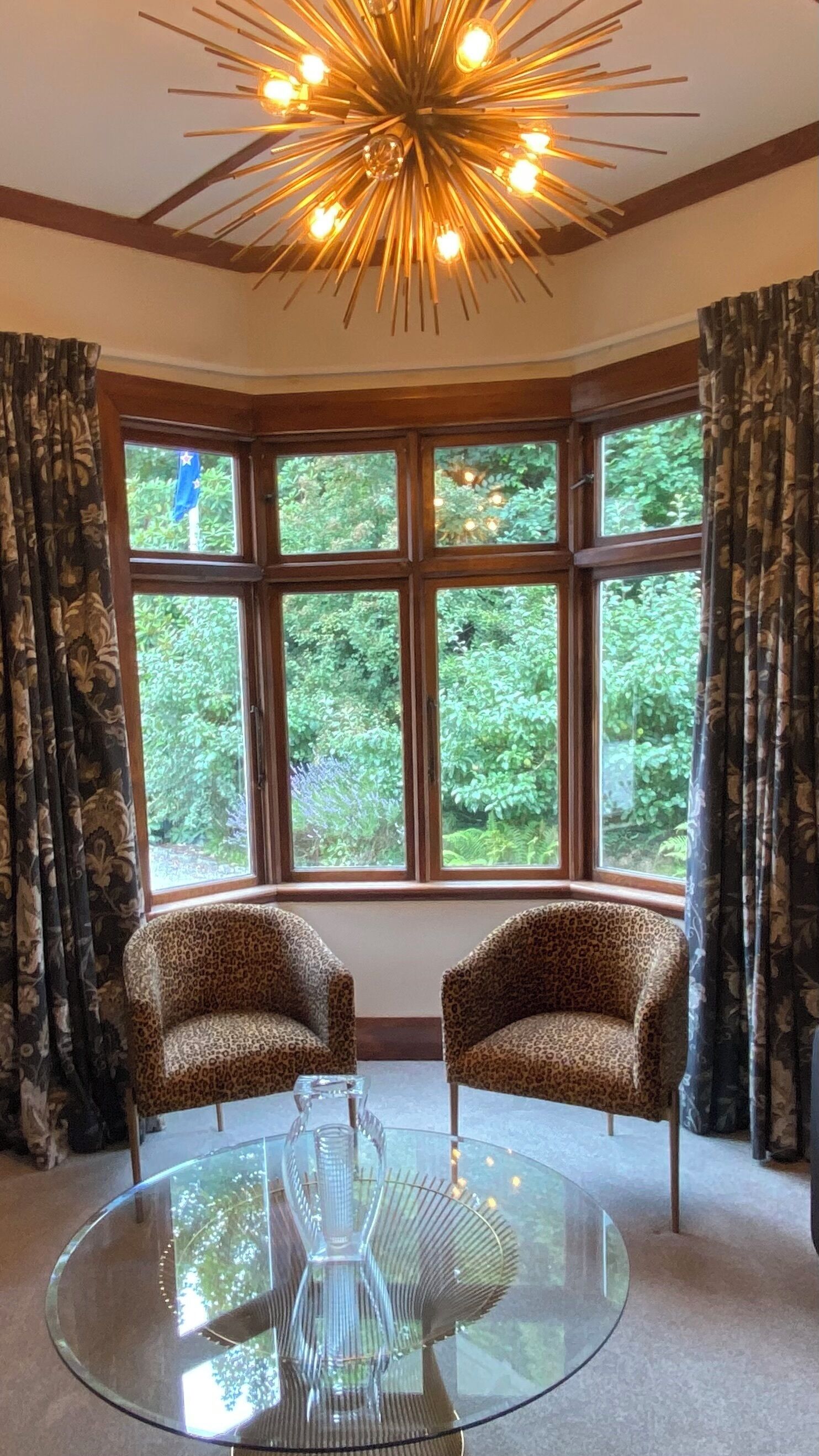 A room with a glass table, two leopard print chairs, a modern ceiling light, and large windows with curtains.