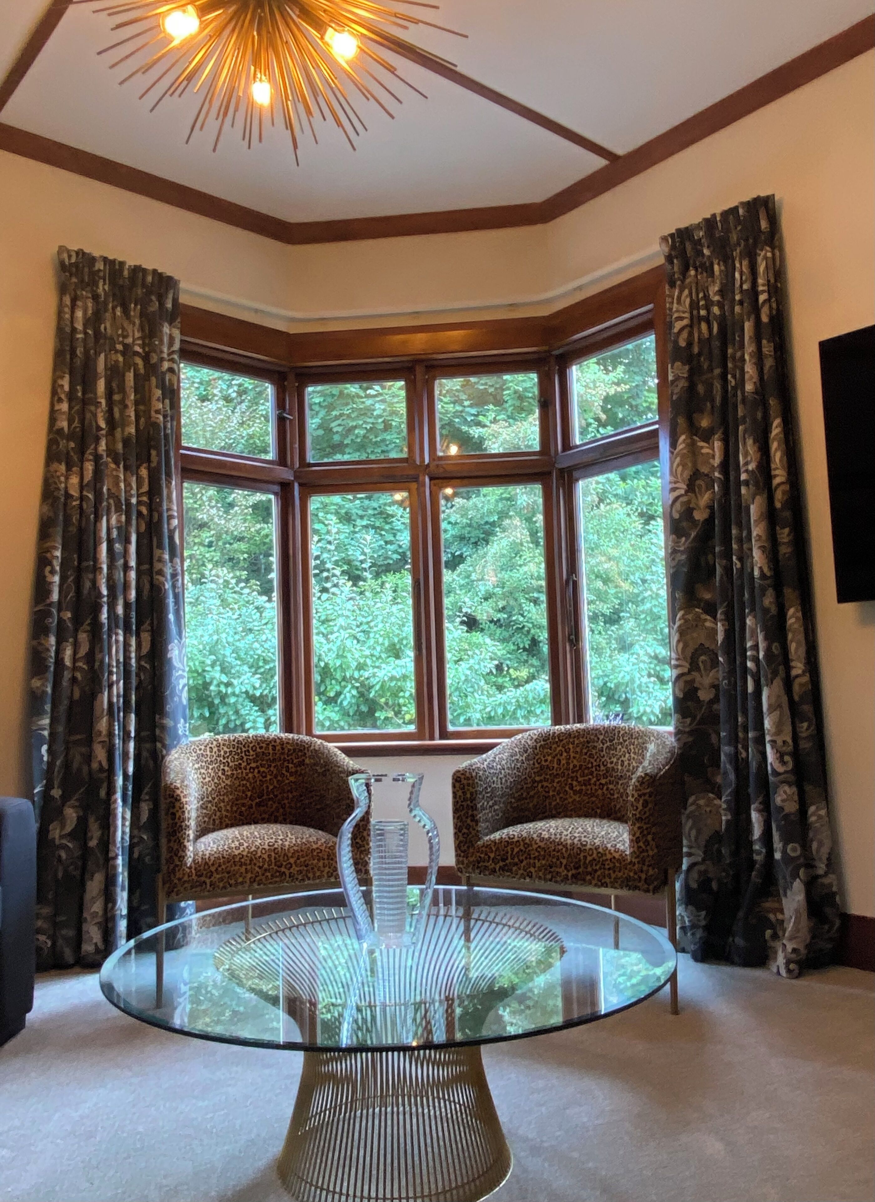 Um quarto com mesa de vidro, duas cadeiras de estampa de leopardo, um amplo parapeito com cortinas e um lustre no teto.
