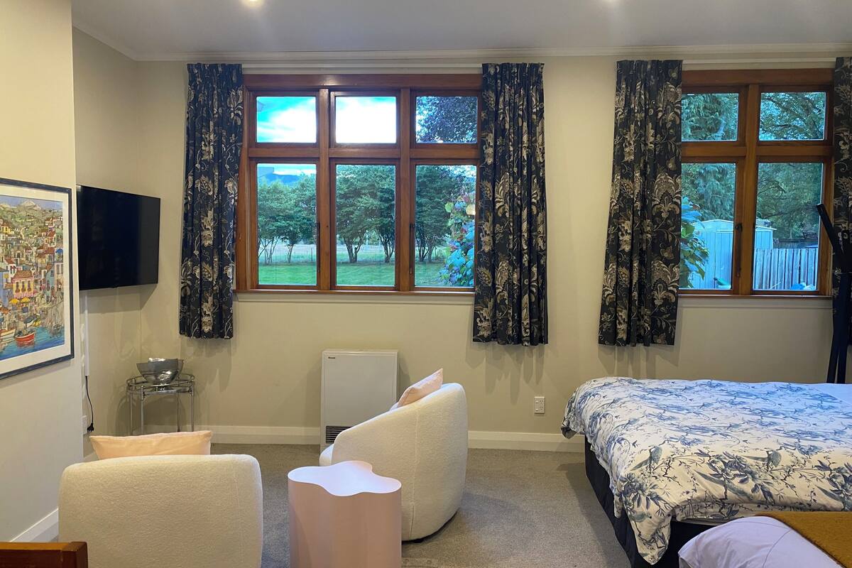 A bedroom with a bed, a chair, a small table, a radiator, and a window with curtains.
