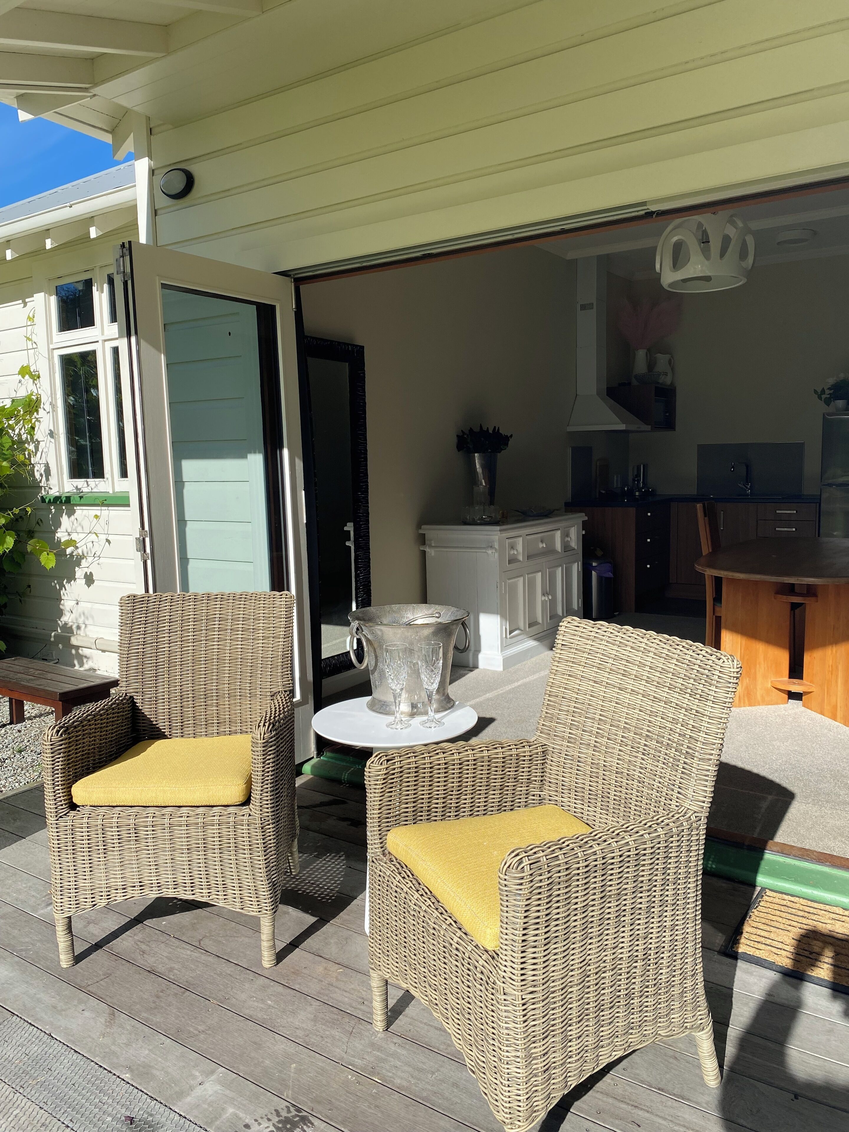Ein überdachter Außenbereich mit Korbstühlen und einem Tisch, eine durchgehende offene Tür zur Küche und ein Blumentopf auf der Terrasse.