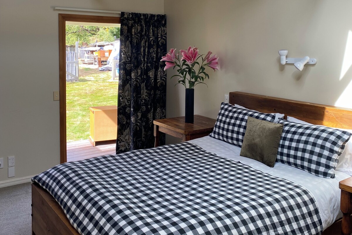 Un dormitorio con edredón a cuadros, cabecera de madera, mesita de noche con un jarrón de flores y vistas al exterior a través de un ventanal.