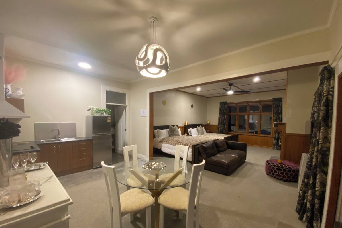 A modern hotel room with a dining area, a bed with a headboard, a sofa, and a large window.