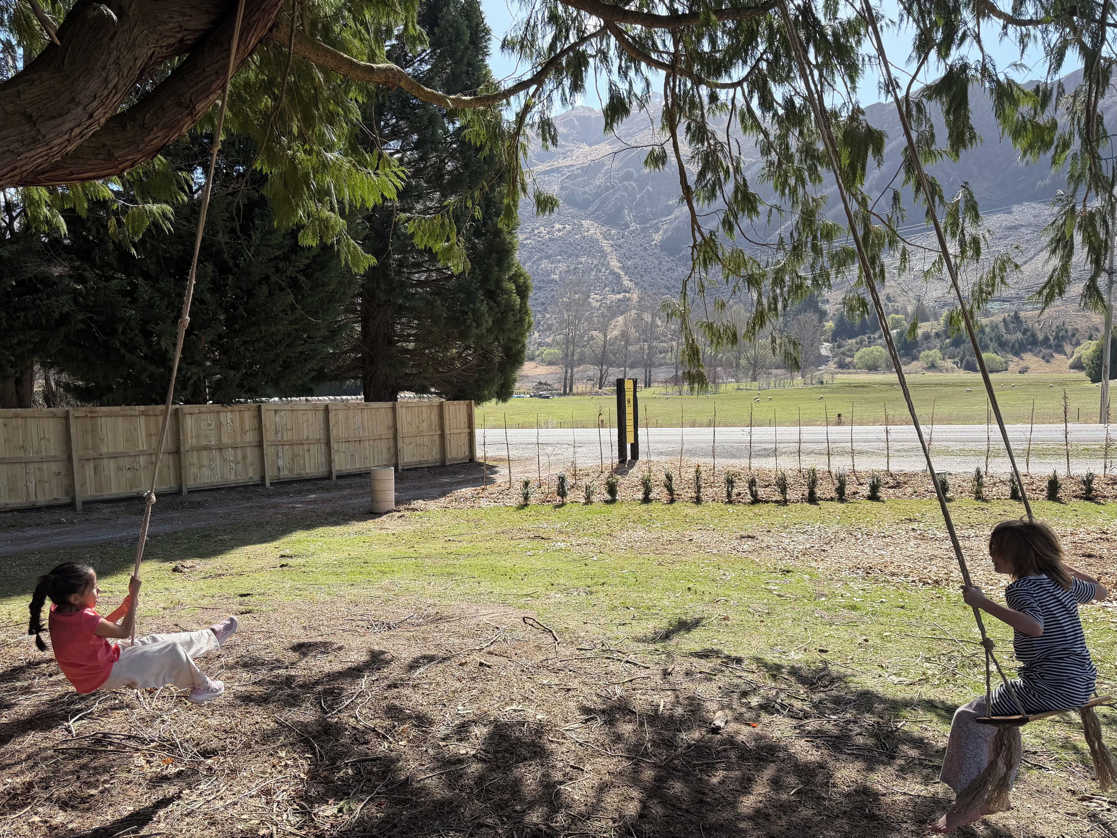 Duas crianças brincando em balanços em um parque, cercado por uma grade de madeira, com vista para montanhas ao longe.