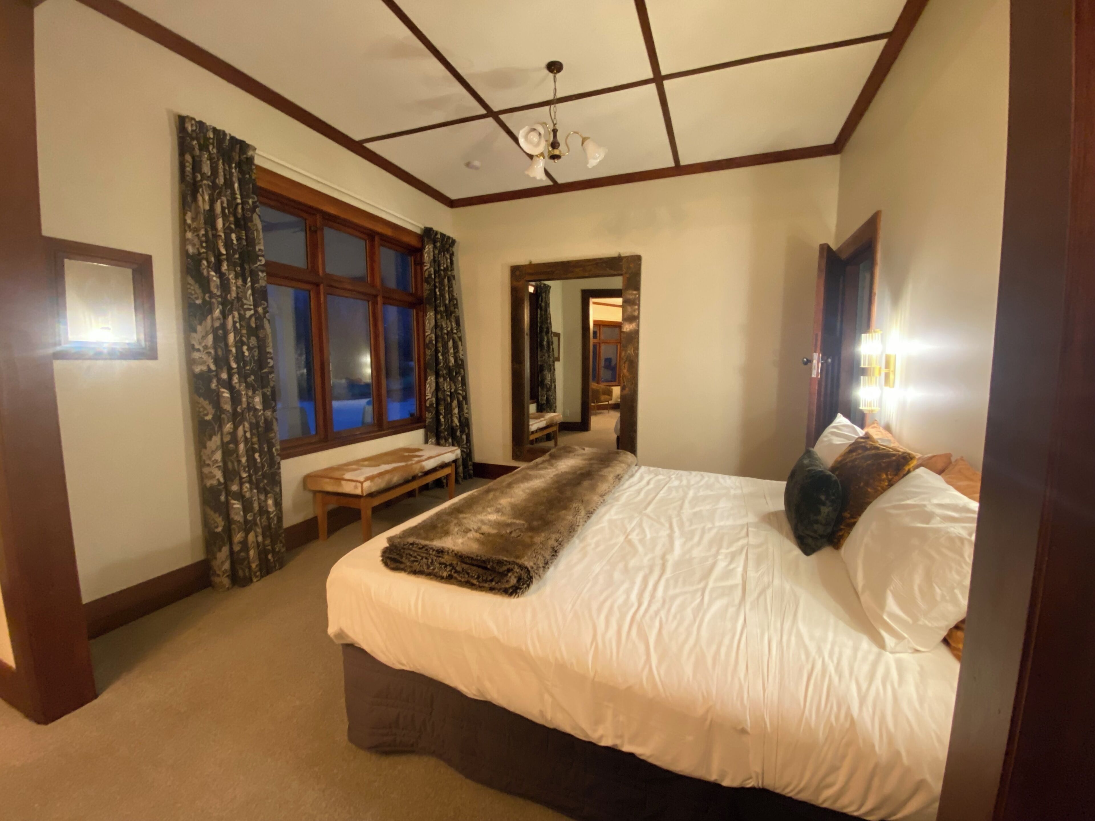 A bedroom with a large bed, a bench, a window with curtains, a ceiling light, and a mirror.
