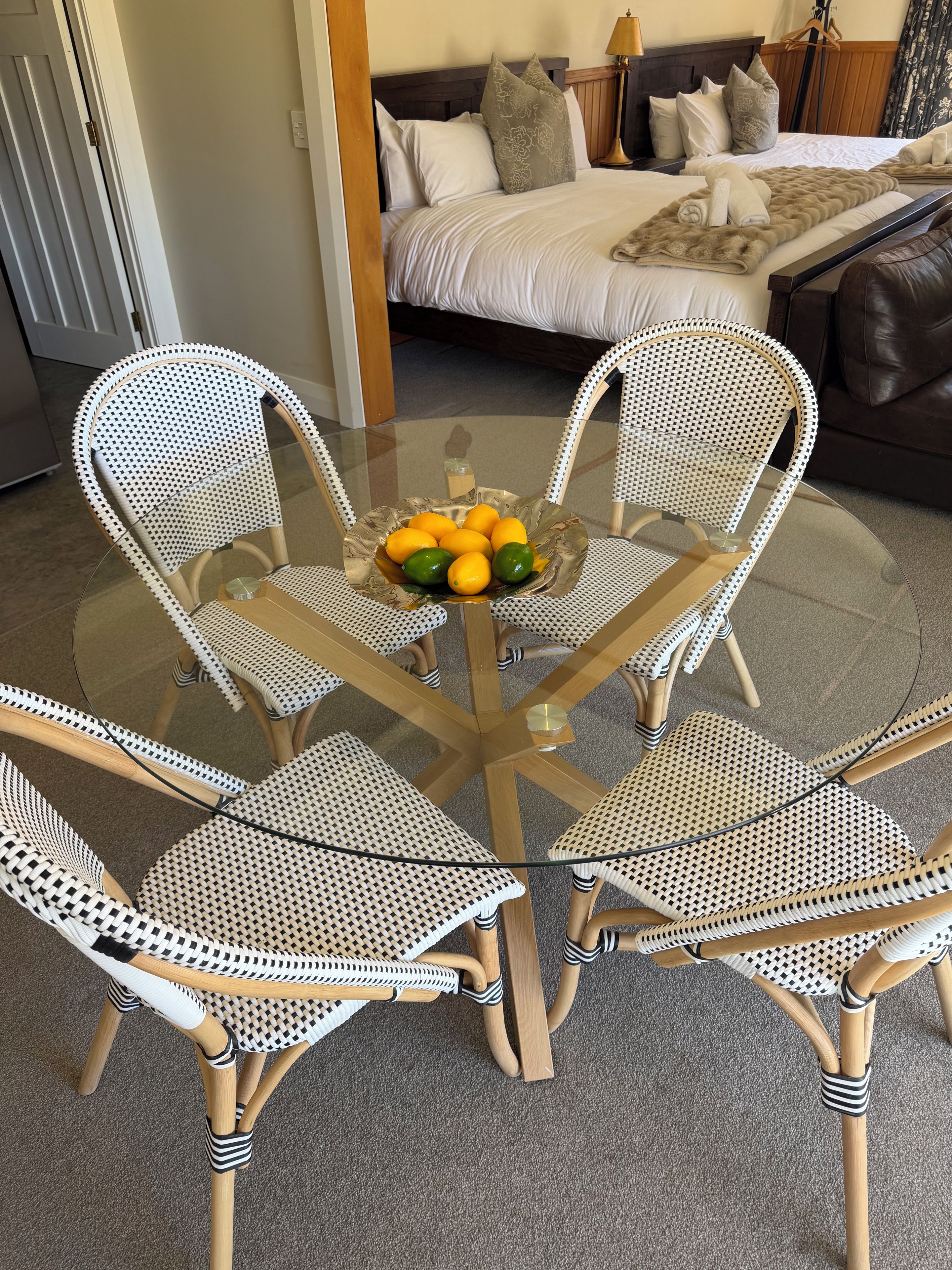 A dining area with a glass table surrounded by four wicker chairs, a bowl of fruit, and a bed with pillows in the background.