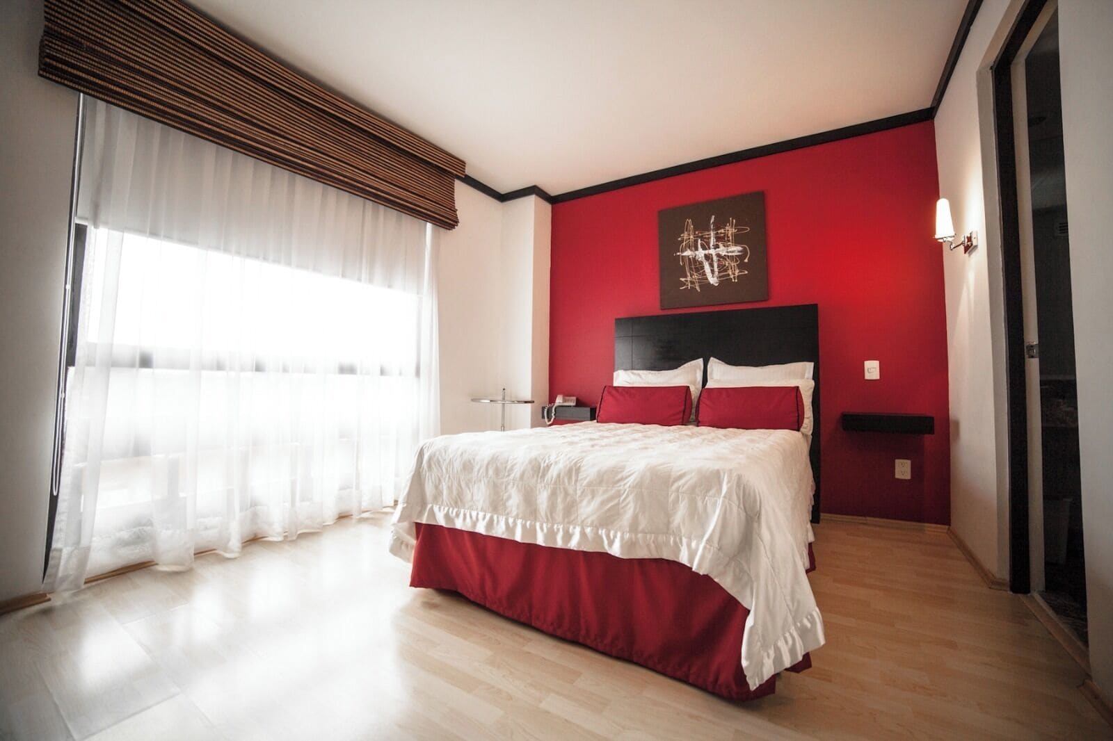 A modern bedroom with a large bed, a red accent wall, and a large window with sheer curtains.