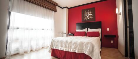 A modern bedroom with a large bed, a red accent wall, and a large window with sheer curtains.