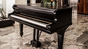 A grand piano in a spacious, well-lit room with a marble floor, a wooden column, and a modern kitchen in the background. - Hotel Inglaterra (Tampico)