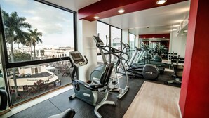 A gym with cardio machines, weight equipment, and a large window offering a view of palm trees and buildings. - Hotel Inglaterra (Tampico)