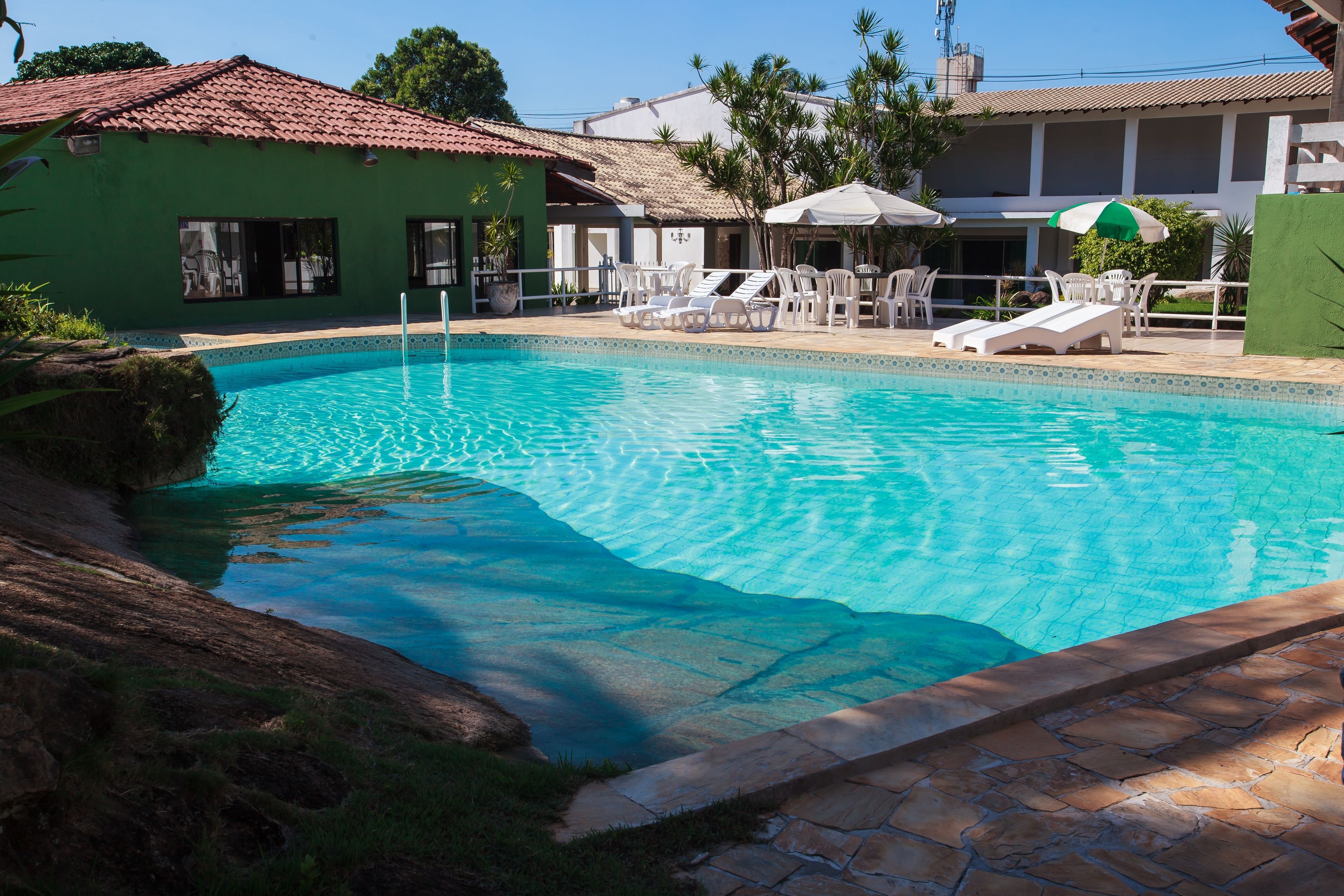 Piscina con sillas de descanso y sombrillas, rodeada por un edificio con techo de tejas y pared verde.